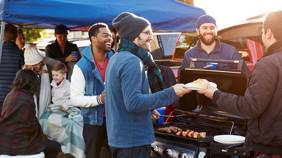 Four smiling men standing in front of a grill full of food while one passes a paper plate with a roll on it  to another. They are next to a blue tent full of men, women and children.