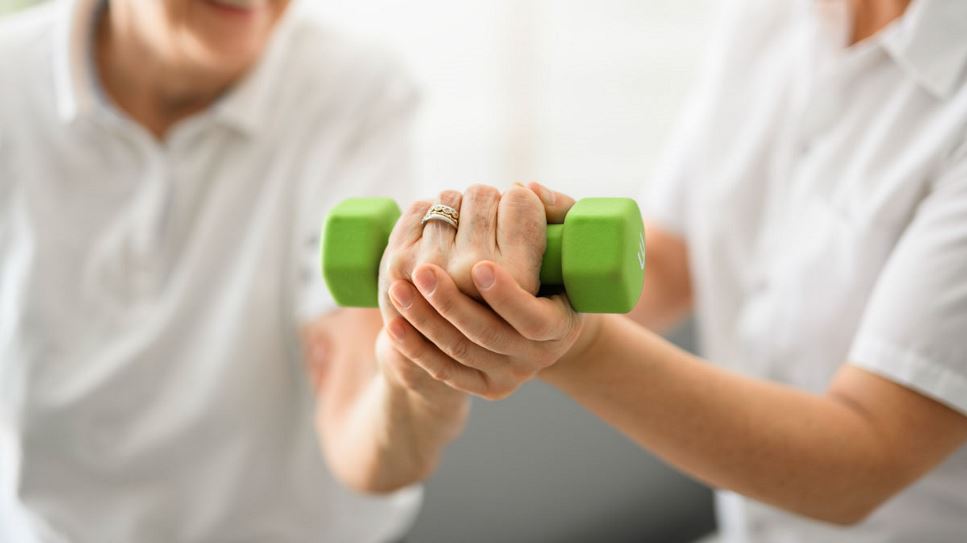An older adult works on lifting a free weight to build muscle strength, with the assistance of a health care provider.